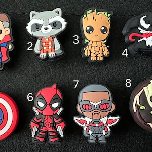 May include: A collection of eight cartoon character charms. The charms include Thor, Rocket Raccoon, Groot, Venom, Captain America's shield, Deadpool, Falcon, and a Groot-themed charm. Each charm is brightly colored and features detailed designs.