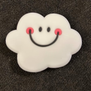 Puede incluir: Un accesorio blanco con forma de nube con una cara sonriente. La nube tiene una boca negra curvada, dos ojos de puntos negros y dos c&iacute;rculos rosados. El accesorio est&aacute; sobre una superficie texturizada gris oscuro.