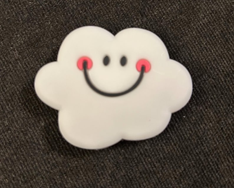 Puede incluir: Un accesorio blanco con forma de nube con una cara sonriente. La nube tiene una boca negra curvada, dos ojos de puntos negros y dos c&iacute;rculos rosados. El accesorio est&aacute; sobre una superficie texturizada gris oscuro.