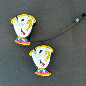 May include: Two cartoon-style cup charms with smiling faces, yellow question mark handles, and colorful trim. The charms are white with black outlines and are connected by a black strap.