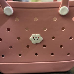 Puede incluir: Un bolso tote rosa con un charm en forma de nube. El bolso tiene un dise&ntilde;o perforado y fijaciones blancas para las asas. El charm de nube tiene una cara sonriente. El bolso est&aacute; hecho de un material duradero.