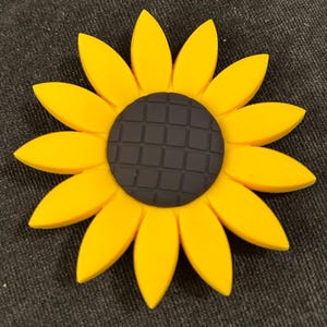 May include: A bright yellow sunflower with a dark gray center. The petals are a vibrant yellow, and the center has a textured, grid-like pattern. The sunflower is a cheerful, decorative item.