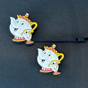 May include: Two cartoon-style, light gray teapots with yellow handles and accents, and a small teacup attached. The teapots have pink and yellow crowns and are set against a dark background. The teapots are approximately 2 inches in height.