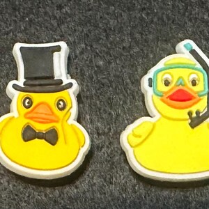 May include: Four yellow rubber duck charms with white borders. One duck holds a rainbow flag, another wears a top hat and bow tie, a third has a snorkel and goggles, and the last holds a purple umbrella.