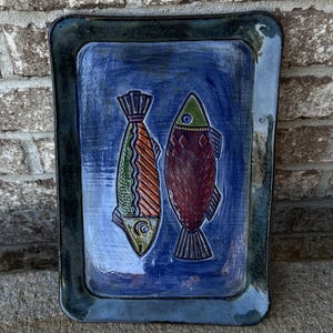 May include: A rectangular ceramic platter with a glossy blue glaze featuring two stylized fish. The left fish has green and orange striped patterns, while the right fish is dark red with a diamond scale texture and a green head.