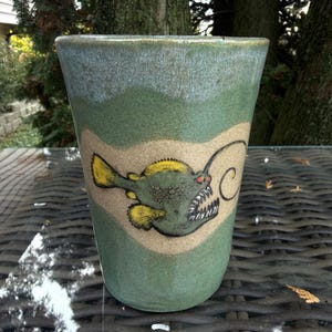 May include: A tapered ceramic tumbler with a sage green glaze and a lighter, speckled beige band around the center. The band features a hand-painted illustration of an anglerfish with yellow fins, sharp white teeth, and a glowing lure.