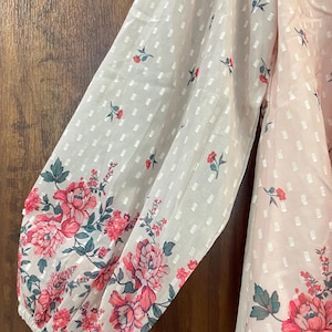 May include: A light pink blouse with long sleeves. The sleeves have a white dotted pattern and are adorned with a floral design in shades of pink and green. The blouse is made of a sheer, lightweight fabric.