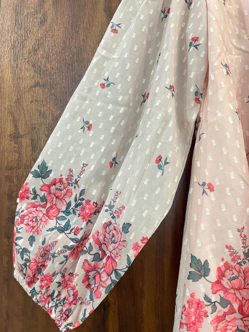 May include: A light pink blouse with long sleeves. The sleeves have a white dotted pattern and are adorned with a floral design in shades of pink and green. The blouse is made of a sheer, lightweight fabric.