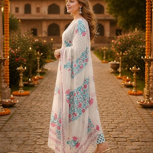 Pu&ograve; includere: Una donna indossa un completo bianco con un motivo floreale turchese e rosa. L'outfit include una lunga sciarpa fluida e pantaloni abbinati. Lo sfondo presenta un edificio ornato e un sentiero in pietra.
