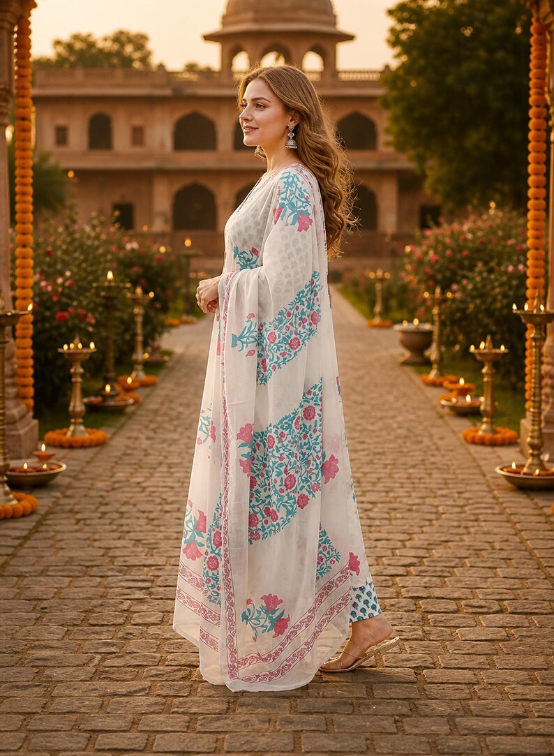 Pu&ograve; includere: Una donna indossa un completo bianco con un motivo floreale turchese e rosa. L'outfit include una lunga sciarpa fluida e pantaloni abbinati. Lo sfondo presenta un edificio ornato e un sentiero in pietra.