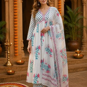 Pu&ograve; includere: Un completo indiano bianco con stampa floreale rosa e turchese. Il completo comprende una tunica a maniche lunghe, pantaloni abbinati e una sciarpa trasparente. L'outfit &egrave; indossato da una persona davanti a uno sfondo tradizionale indiano.