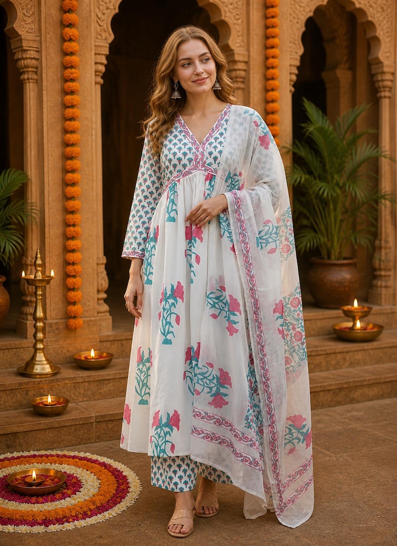 Pu&ograve; includere: Un completo indiano bianco con stampa floreale rosa e turchese. Il completo comprende una tunica a maniche lunghe, pantaloni abbinati e una sciarpa trasparente. L'outfit &egrave; indossato da una persona davanti a uno sfondo tradizionale indiano.