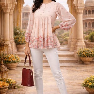 May include: A light pink top with floral detailing and long sleeves, paired with white pants. The top has a round neckline and a peplum-style hem. A burgundy handbag is held in the hand. The background features an ornate architectural setting.