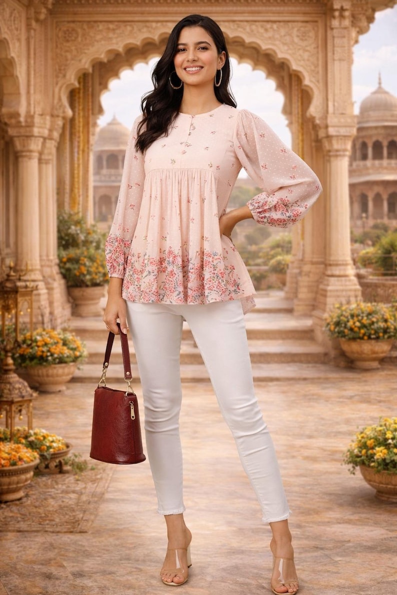 May include: A light pink top with floral detailing and long sleeves, paired with white pants. The top has a round neckline and a peplum-style hem. A burgundy handbag is held in the hand. The background features an ornate architectural setting.