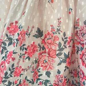 May include: Close-up of a light pink fabric with a white dotted pattern. The bottom edge features a floral border with large pink peonies, smaller pink flowers, and green leaves. The fabric has a delicate, sheer quality.