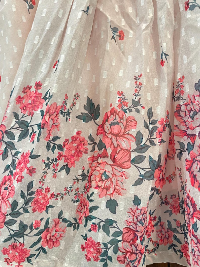 May include: Close-up of a light pink fabric with a white dotted pattern. The bottom edge features a floral border with large pink peonies, smaller pink flowers, and green leaves. The fabric has a delicate, sheer quality.