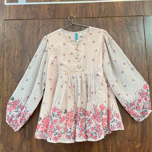 May include: A light pink blouse with long sleeves and a floral pattern. The blouse features a round neckline, button details, and a gathered waist. The sleeves and hem are adorned with a vibrant pink floral border.