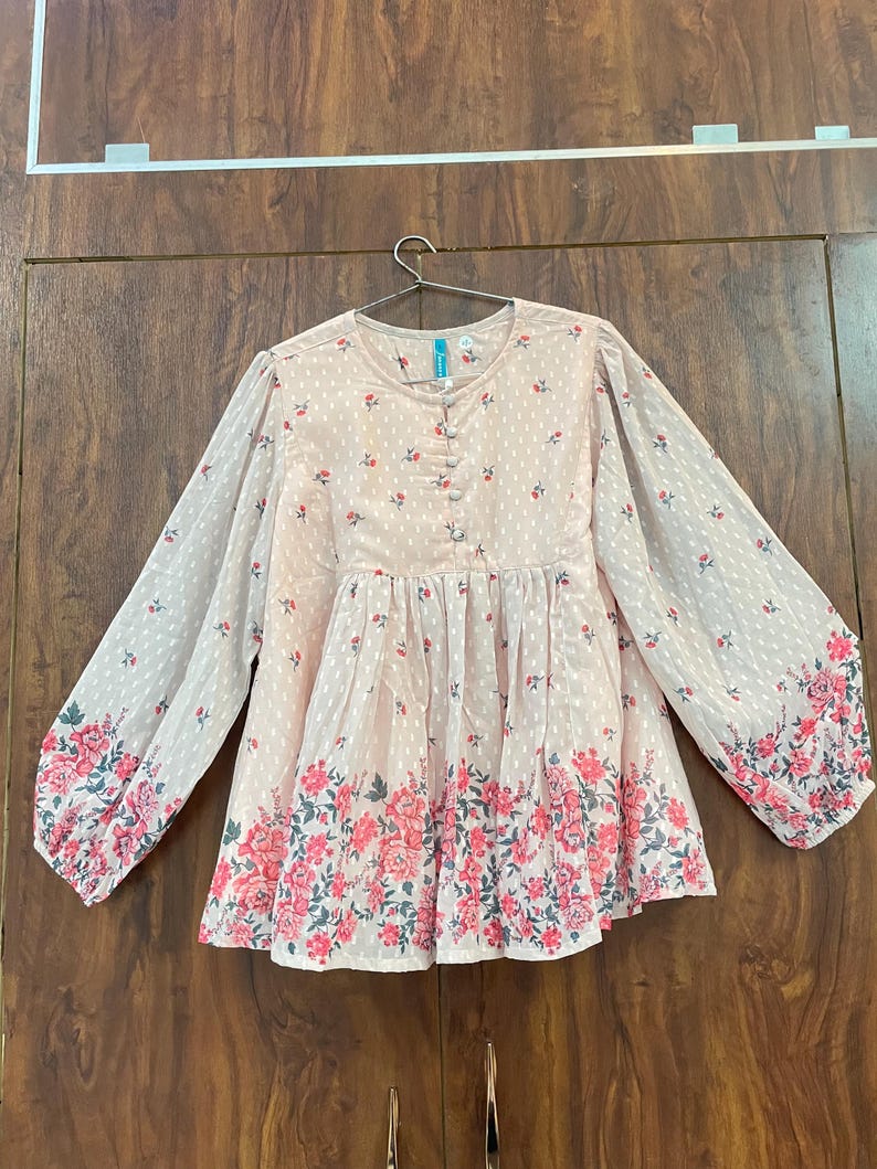 May include: A light pink blouse with long sleeves and a floral pattern. The blouse features a round neckline, button details, and a gathered waist. The sleeves and hem are adorned with a vibrant pink floral border.
