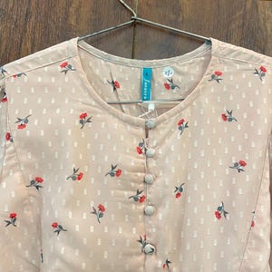 May include: A light pink blouse with a floral pattern of red flowers and green leaves. The blouse has a round neckline, a button-down front, and a gathered waist. The brand name "Janasya" is visible on the tag.