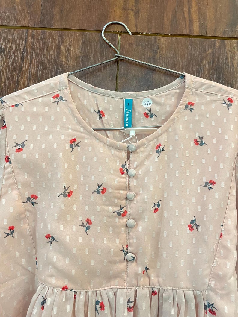 May include: A light pink blouse with a floral pattern of red flowers and green leaves. The blouse has a round neckline, a button-down front, and a gathered waist. The brand name "Janasya" is visible on the tag.