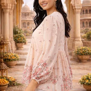 May include: A light pink blouse with a floral pattern on the sleeves and hem. The blouse has long sleeves and a gathered waist. The background features an ornate architectural setting with arches and domes, and potted plants.