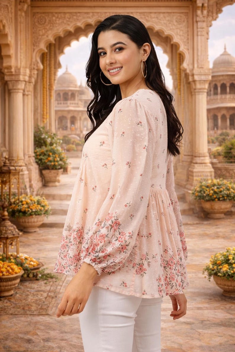 May include: A light pink blouse with a floral pattern on the sleeves and hem. The blouse has long sleeves and a gathered waist. The background features an ornate architectural setting with arches and domes, and potted plants.