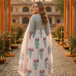 Pu&ograve; includere: Una donna indossa un abito indiano bianco con stampa floreale e una sciarpa trasparente abbinata. L'abito presenta un corpetto con un piccolo motivo verde e bianco e una gonna con un disegno floreale rosa e verde. Lo sfondo include un sentiero di pietra e un edificio.