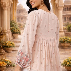 May include: A light pink blouse with long sleeves and a floral pattern. The sleeves have a floral design at the cuffs. The blouse has a gathered waist and a V-neck. The model is wearing white pants.