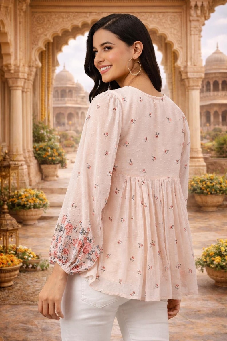May include: A light pink blouse with long sleeves and a floral pattern. The sleeves have a floral design at the cuffs. The blouse has a gathered waist and a V-neck. The model is wearing white pants.