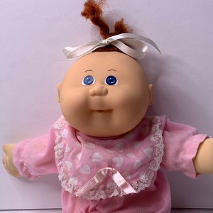 May include: A Cabbage Patch Kid doll with brown hair, blue eyes, and a pink outfit with white hearts. The doll is sitting with its legs crossed and has a white bow in its hair.