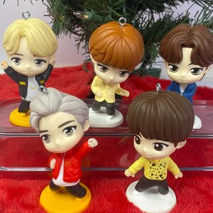 May include: Five miniature figurines with large eyes and stylized hair, each wearing different outfits. The figurines are on yellow and white bases, and are arranged on a red, fuzzy surface, with a green Christmas tree in the background.