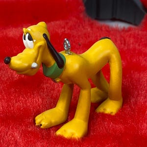 May include: A yellow Pluto figurine with a black ear, green collar, and black nose. The Disney character is posed on all fours, with a silver loop for hanging. The ornament is set against a red, fuzzy background.