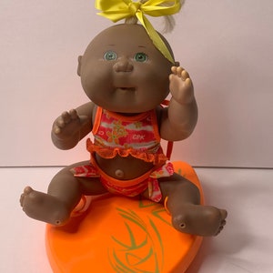 May include: A Cabbage Patch Kids doll with dark skin, green eyes, and blonde hair tied with a yellow bow. The doll wears a swimsuit and sits on an orange surfboard with green designs.