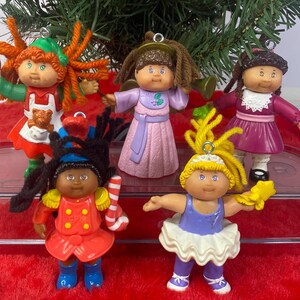 May include: A collection of five Cabbage Patch Kids Christmas ornaments. Each doll is dressed in a unique outfit, including a chef, a ballerina, and a soldier. The ornaments are made of plastic and have small metal loops for hanging.