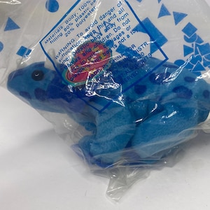 Może przedstawiać: Niebieska pluszowa żaba z ciemniejszymi niebieskimi plamkami, zapakowana w przezroczystą plastikową torbę. Żaba wykonana jest z akrylu i poliestru. Torba posiada etykietę ostrzegawczą o niebezpieczeństwie uduszenia.