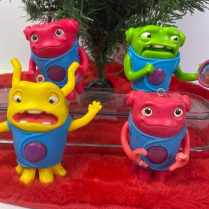 Puede incluir: Cuatro adornos de personajes coloridos. Los adornos son en tonos de rosa, verde y amarillo, cada uno con un chaleco azul y un botón morado. Cuelgan de un árbol de Navidad.