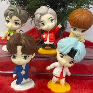 May include: Five small, cartoon figurines with varied hairstyles and outfits, each on a small yellow base. The figurines are arranged on a red, fuzzy surface, with a Christmas tree in the background. Each figure has a silver loop for hanging.