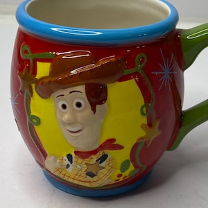 Puede incluir: Una taza de cer&aacute;mica con una imagen de dibujos animados de Woody de Toy Story. La taza tiene un exterior rojo con un marco ovalado amarillo alrededor de la cara de Woody. El asa es verde y el borde es azul.