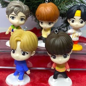 May include: Five small, cartoon-style figurines designed as Christmas ornaments, each with a unique hairstyle and outfit. The figures are on white bases, set against a Christmas tree and a red, fuzzy surface. Each figure has a loop for hanging.
