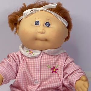 May include: A vintage Cabbage Patch Kids doll with red hair and blue eyes. The doll is dressed in a pink and white gingham pajama set, featuring floral and butterfly embroidery. A white ribbon headband is tied around the doll's hair.