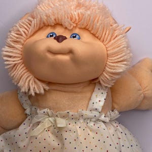 May include: A vintage Cabbage Patch Kids doll with peach-colored yarn hair and blue eyes. The doll wears a white dress with colorful polka dots and a bow. The doll's face has a smiling expression.