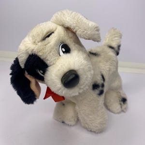 Puede incluir: Un peluche de cachorro dálmata con cuerpo crema, manchas negras y una oreja negra. Tiene una pajarita roja y una nariz negra. El cachorro está sobre una superficie blanca.