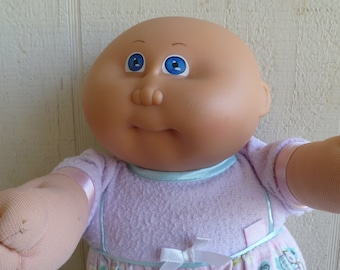 Puede incluir: Una muñeca Cabbage Patch Kid con un vestido lavanda y rosa con un lazo azul. La muñeca tiene la cabeza calva, ojos azules y una tez rosada.