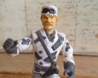 May include: A white and gray action figure with a white hat and goggles, a black belt, and a gray and white camouflage pattern. The figure has a mustache and a light brown beard.