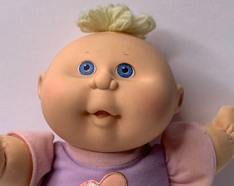 Puede incluir: Una muñeca Cabbage Patch Kid con cabello rubio y ojos azules que lleva un atuendo morado y rosa con un diseño en forma de corazón.