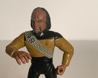 May include: A plastic action figure of a Star Trek character, Worf, in his Starfleet uniform. He has a dark brown skin tone, black hair, and a black and gold uniform with a silver belt. He is in a fighting pose with his arms raised.
