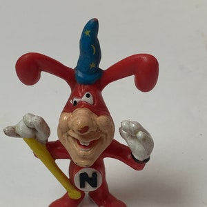 May include: A red and white toy figure of a cartoon character wearing a blue wizard hat with stars. The character has a large nose, a wide smile, and is holding a yellow stick. The character is wearing a red suit with a large black letter "N" on the chest.