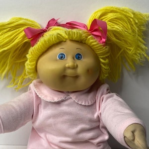 Könnte beinhalten: Eine Cabbage Patch Kid Puppe mit blonden Garnhaaren in Zöpfen mit rosa Schleifen. Die Puppe trägt ein rosa Langarmshirt mit weißem Kragen und weiße Hosen mit rosa Streifenmuster.