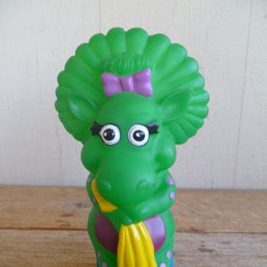 Może przedstawiać: Zielona plastikowa zabawka przedstawiająca postać z fioletową kokardką, żółtym szalikiem i fioletowymi butami. Postać ma duże oczy i zielone, kręcone włosy.
