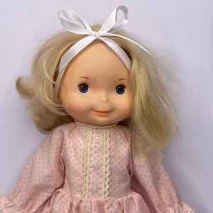 May include: A vintage doll with blonde hair and blue eyes. The doll is dressed in a pink polka dot dress with lace trim and a white bow headband. The dress has long sleeves and a frilled hem.